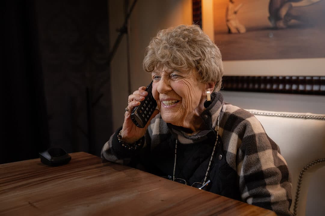 Senior woman in her 70s sitting comfortably at a kitchen table, smiling while talking on a corded home phone.